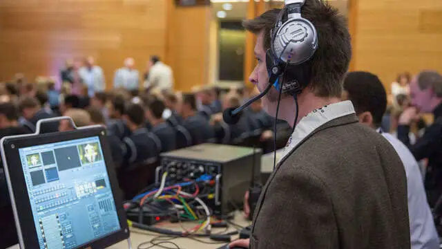 Man with headset operates audio-visual equipment at a crowded sports management event in a conference room.