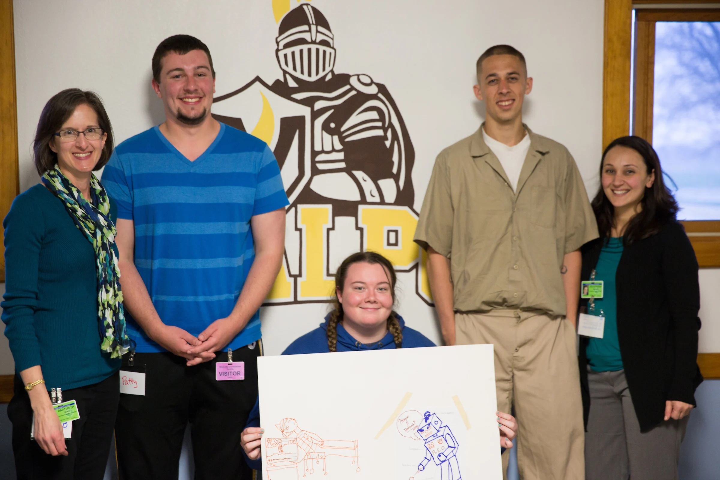 Five people pose and smile; one is seated holding a drawing in front of a knight mascot logo on the wall.