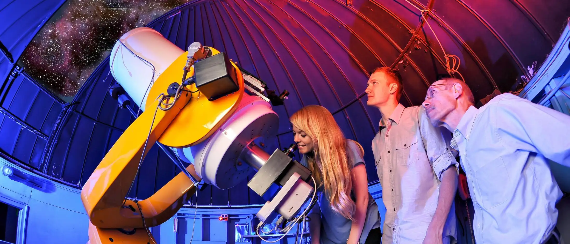 Three people observe the night sky through a large yellow telescope inside a domed observatory, with stars visible outside.