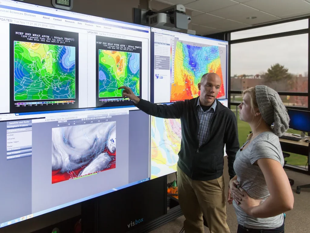 Man points at weather maps on large screens while talking to a woman in an office with more displays.