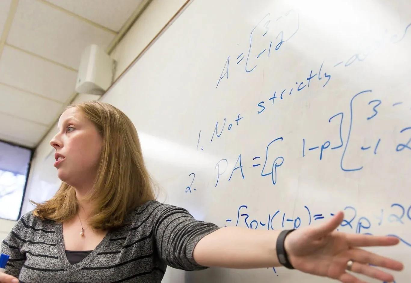 Woman explaining mathematical equations and analytics modeling written on a whiteboard in a classroom setting.