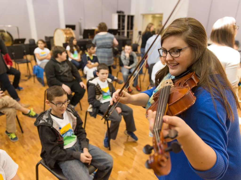 A woman provides music education by playing violin for seated children in a classroom, as the kids watch and listen attentively.