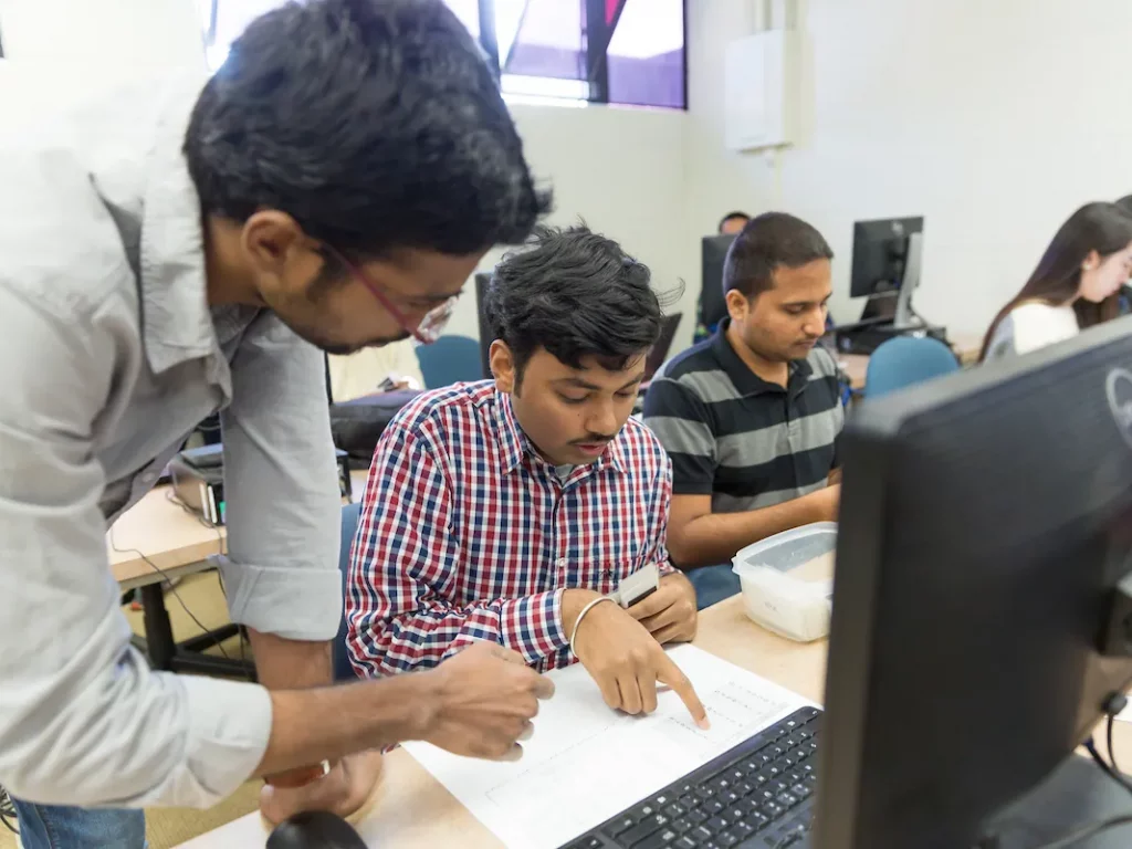 A man helps a student with an Information Technology worksheet in a computer lab, while others work at their desks.