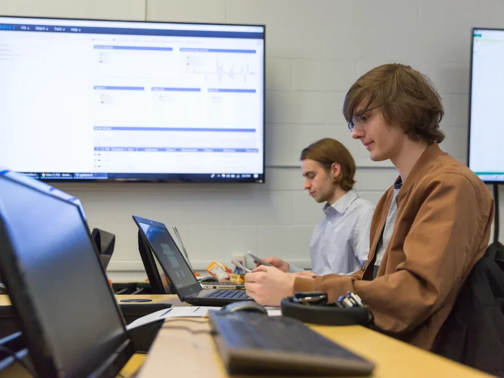 Two people work at laptops in a classroom, exploring Data Science on large screens displaying data behind them. Desks with various electronic devices are also visible.