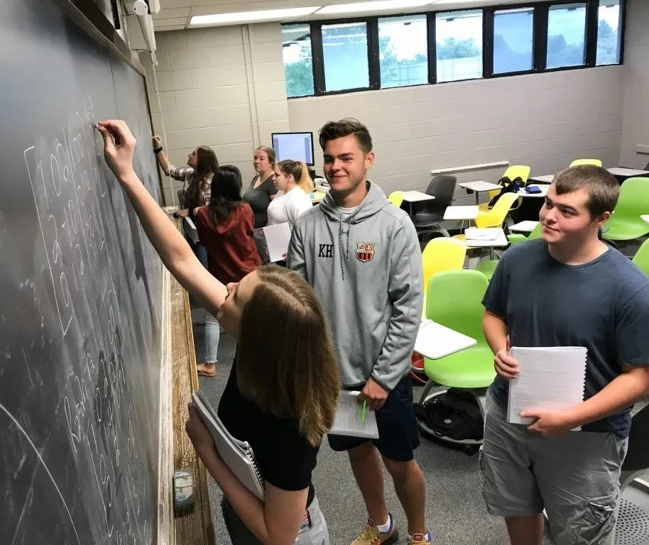 Students write on a chalkboard in a classroom while others watch and hold notebooks.