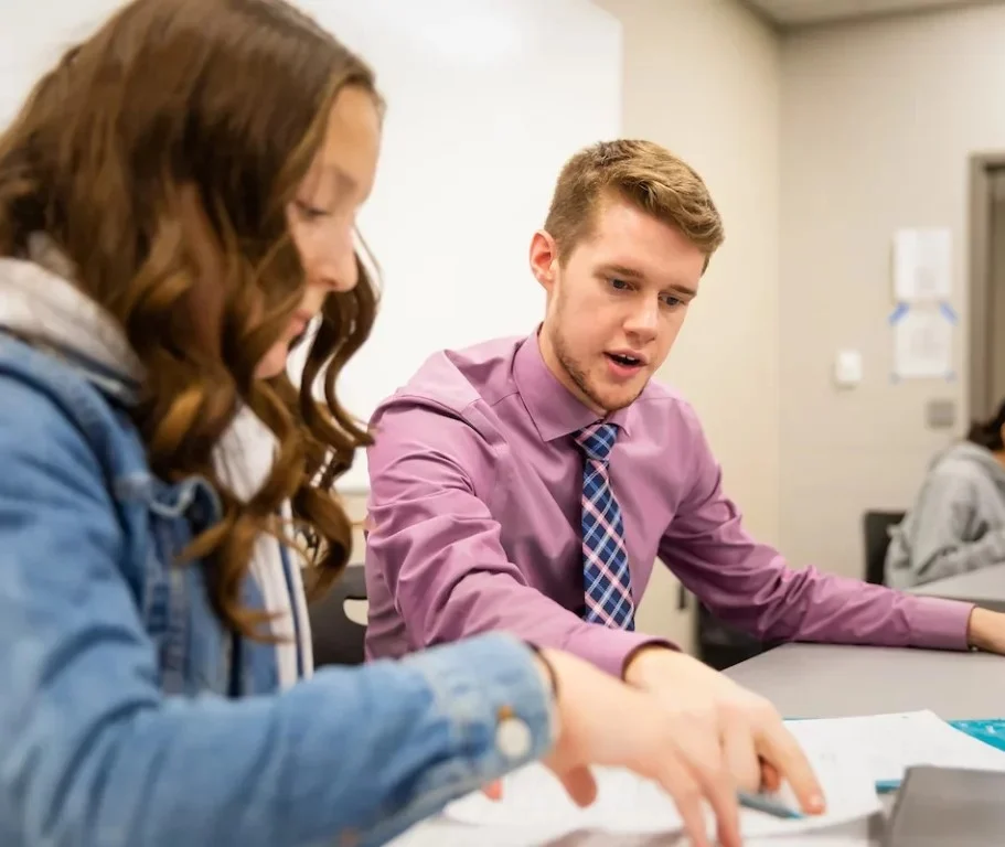 A man in a dress shirt helps a student with schoolwork at a classroom desk, highlighting the importance of elementary education.