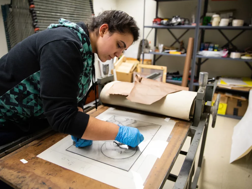 A person wearing blue gloves works on an art drawing at a printmaking press in an art studio.