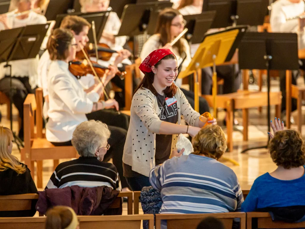 A woman hands something to seated audience members at a concert, where musicians play string instruments in the background, creating a soothing atmosphere of therapeutic music.