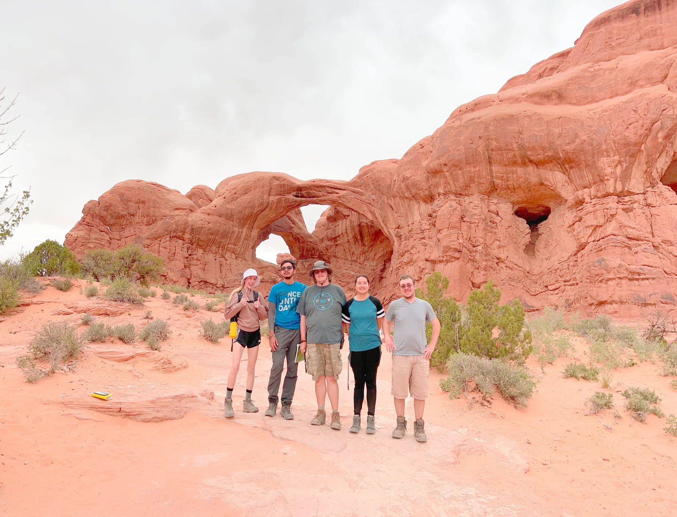 Five people, all Geography BA graduates, stand smiling in front of red rock arches in a desert landscape under a cloudy sky.