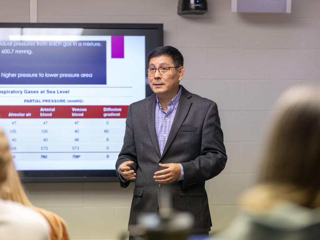 A man in a suit stands in front of a classroom, giving a lecture on Exercise Science with a presentation slide about respiratory gases displayed on the screen behind him.