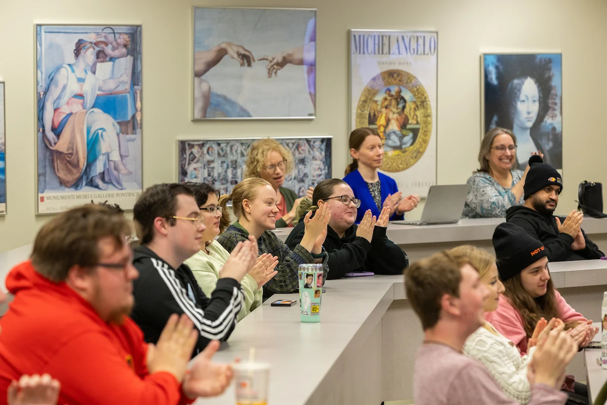 Students sit in an English classroom, smiling and applauding, with art posters visible on the walls behind them.