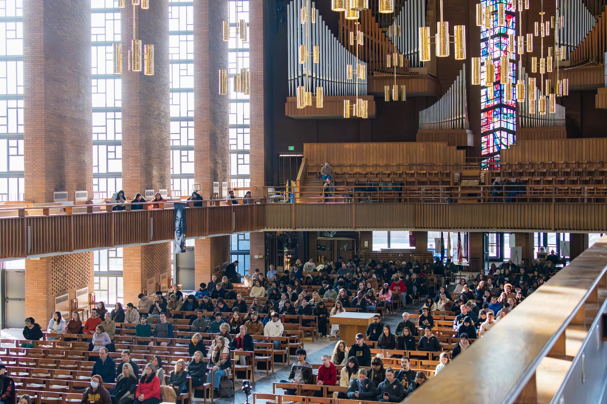 People seated in a large, modern church with stained glass and tall organ pipes, listening attentively—perfect for Religious Studies gatherings or discussions.