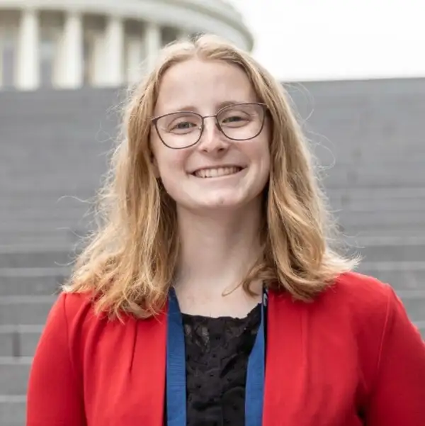 A smiling young woman with glasses and a red jacket stands in front of steps and a domed building, exuding confidence and open communication.