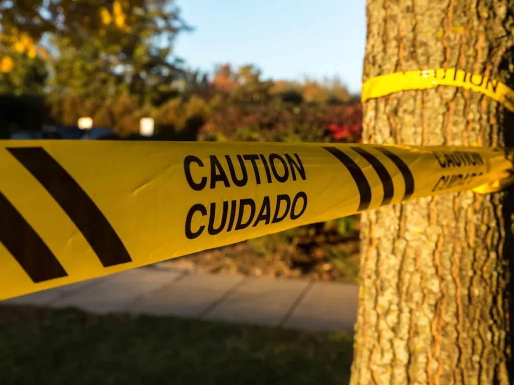 Yellow caution tape in English and Spanish is wrapped around a tree in a sunlit outdoor area.
