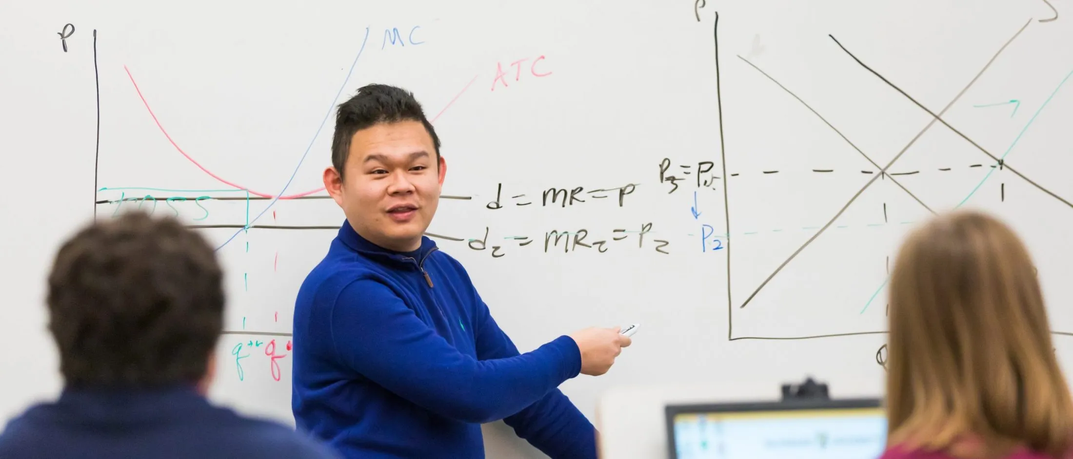 A man in a blue sweater explains economic graphs and equations on a whiteboard to a classroom of students.