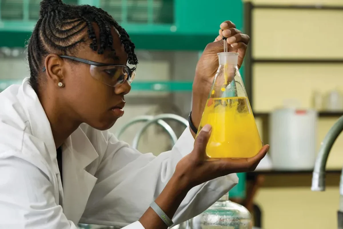 A scientist wearing safety goggles and a lab coat examines a yellow liquid in an Erlenmeyer flask inside a laboratory.