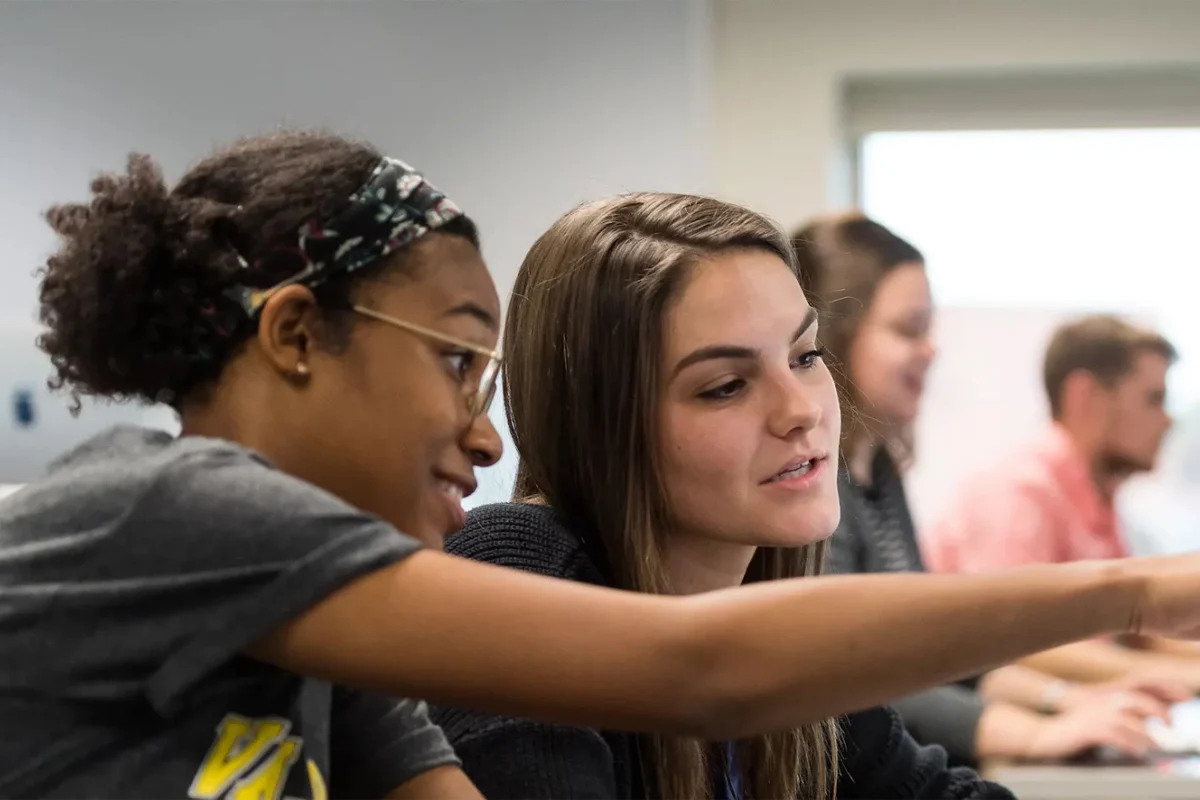 Two students in a classroom collaborate through effective communication, focusing on something on a screen, with others blurred in the background.