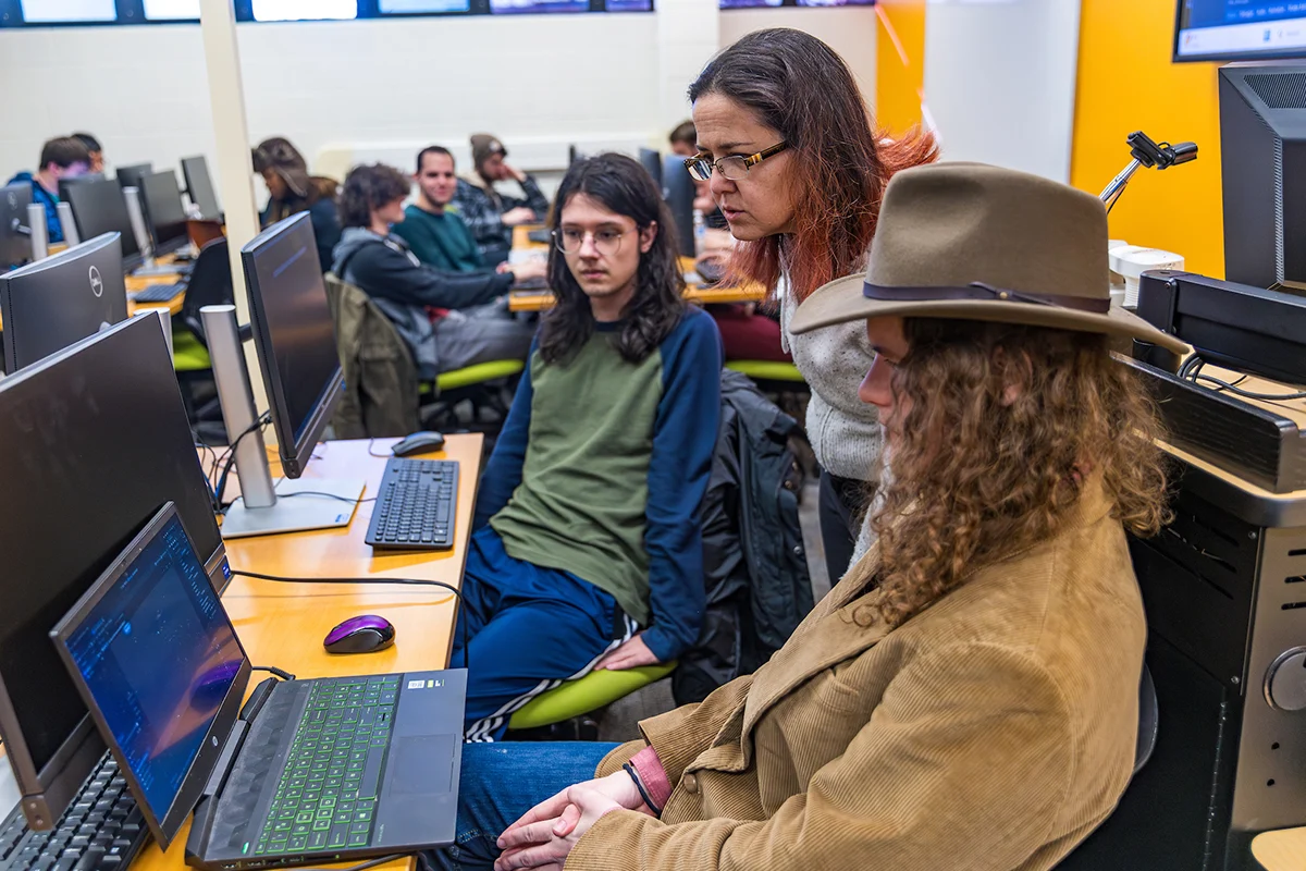 Three people collaborate at a computer in a Computer Science classroom, with others working at computers in the background.