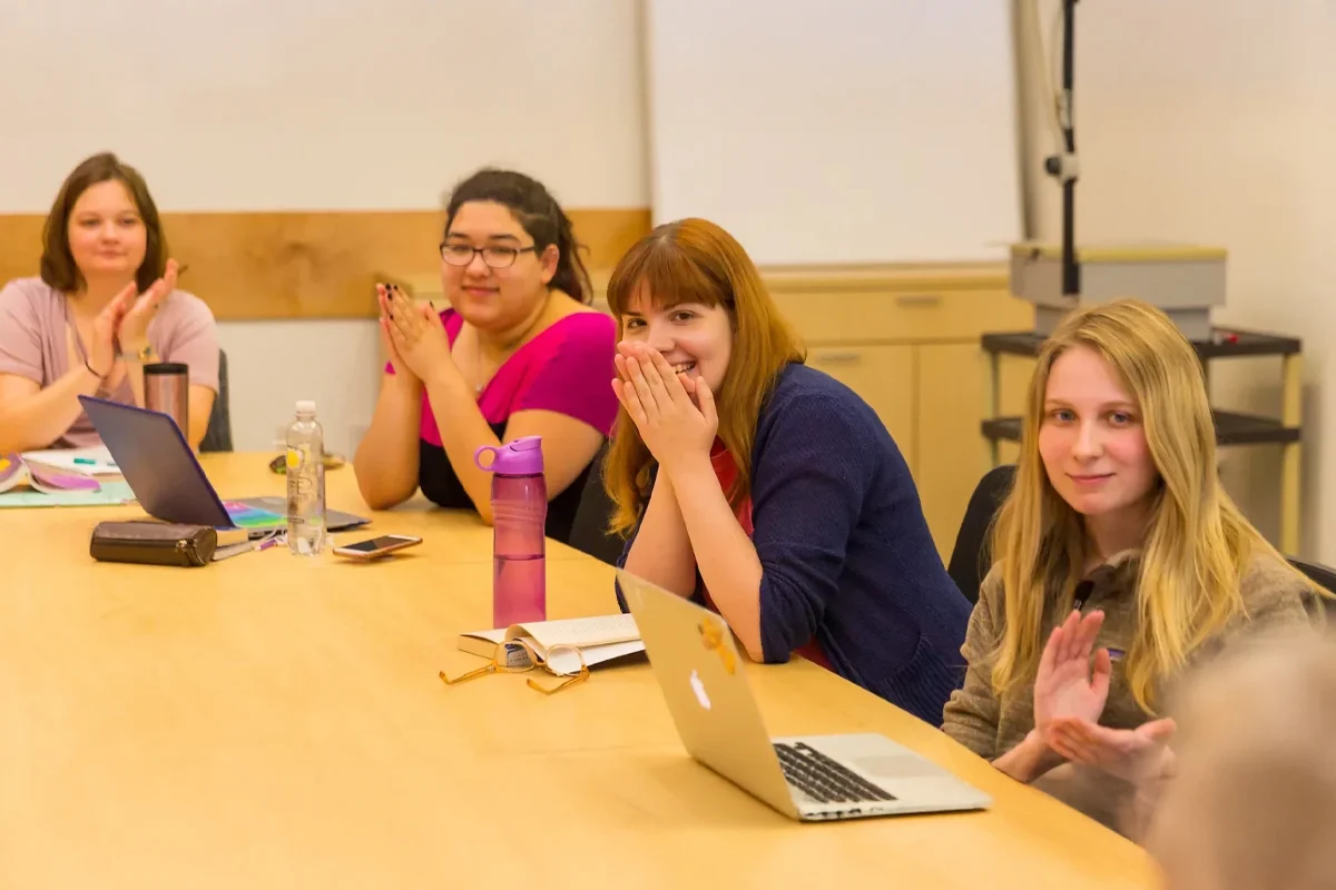 Four women sit around a conference table with laptops and notebooks, looking toward the camera and clapping, in a brightly lit meeting room—celebrating a successful creative writing session.