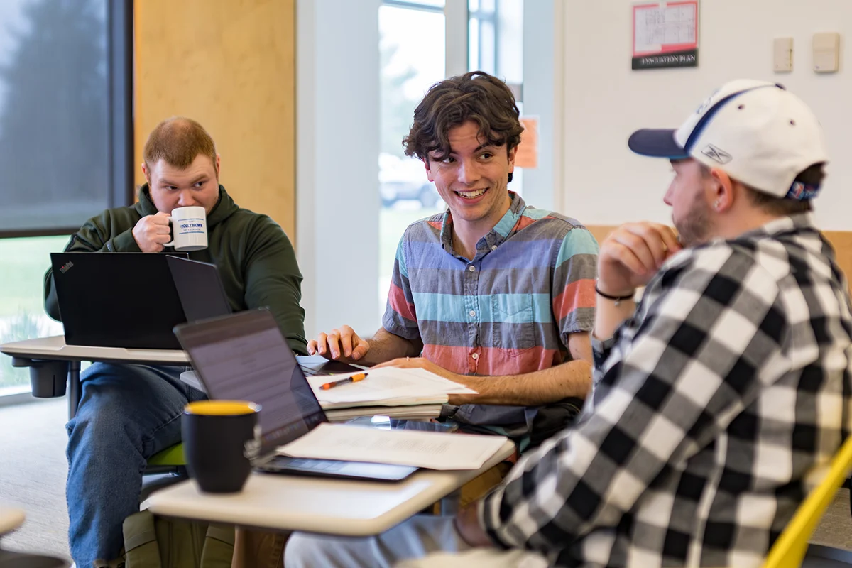 Three young men sit at desks, discussing history while studying with laptops, notebooks, and coffee mugs.