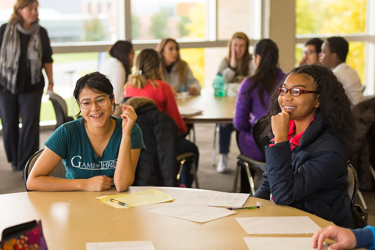 Two students sit smiling at a round table in a bright classroom, with others talking in the background, reflecting the vibrant atmosphere of International Undergraduate Admission.