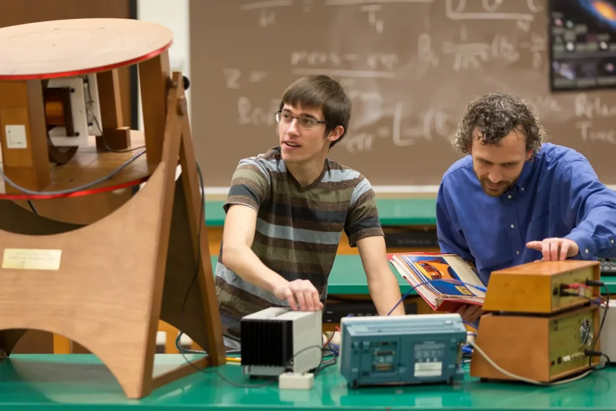 Two men work with scientific equipment in a classroom with a chalkboard in the background.