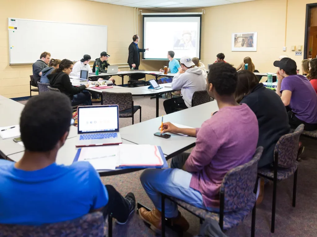 College students sit in a Political Science classroom with laptops, watching a teacher give a presentation at the front.