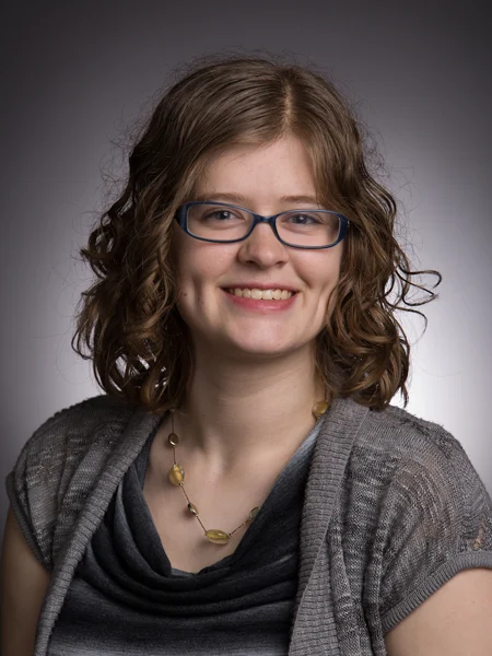 Alumni Samantha Cassell, a young woman with curly brown hair, glasses, and a gray sweater, smiles at the camera.