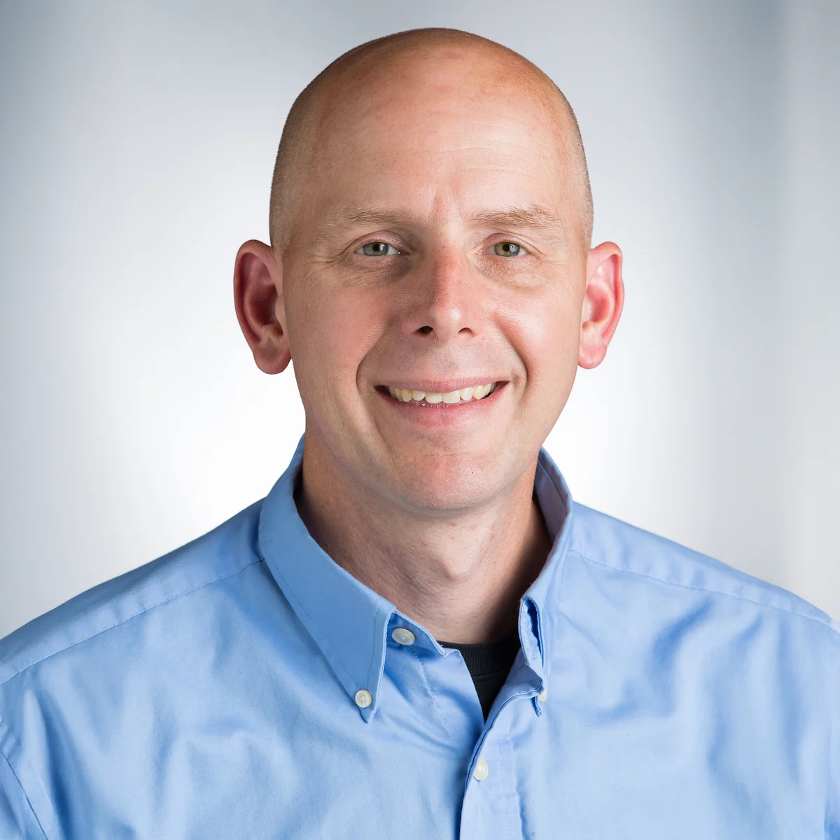 Bald man in a light blue button-up shirt smiling at the camera with a light background.