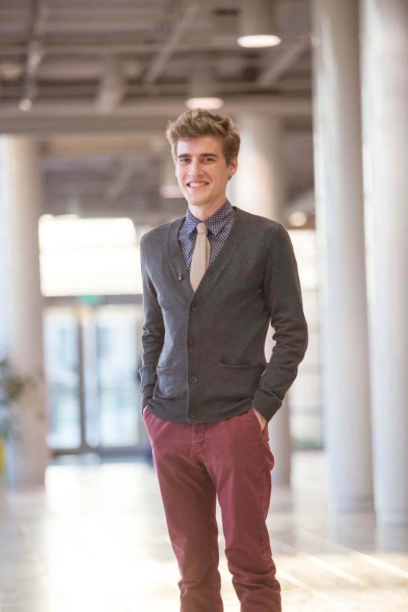 Stefan Roseen, a young man in a cardigan and tie, stands indoors with hands in pockets, smiling at the camera.