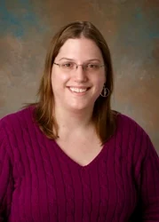 Smiling Valpo Math Professor Lara Pudwell, MAA's Alder Award recipient, with glasses and straight brown hair, wearing a purple sweater and seated in front of a brown background.