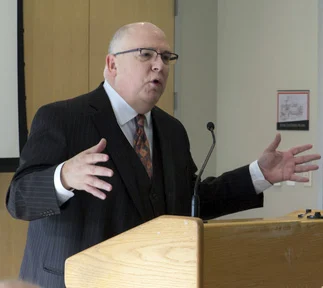 A generous mentor in a suit speaks passionately at a podium, gesturing with open hands.