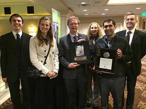 Six members of the solar team in formal attire, two proudly holding awards, stand together and smile in a hotel hallway.