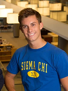 Smiling young man wearing a blue "Sigma Chi" T-shirt stands indoors with a modern background, embodying the spirit of going beyond formulas in everyday life.