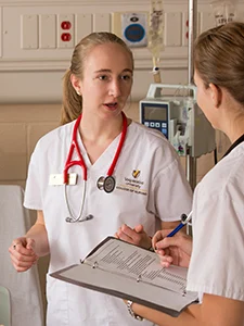 Two healthcare workers in white uniforms discuss their career paths; one holds a clipboard while the other with a stethoscope highlights the dedication to their vocation.