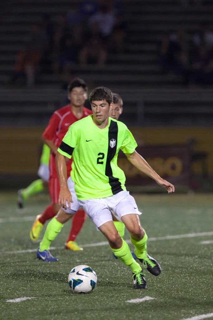 Soccer player Todd Vervynckt in a neon green jersey dribbles the ball on a field, with opponents in red chasing behind him.