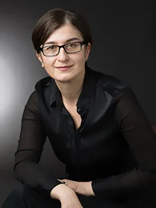 Woman with short brown hair, glasses, and a black blouse sits against a dark background, smiling softly—a portrait that reflects her dedication to academic teamwork and faculty collaboration.
