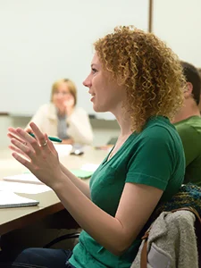 Woman with curly hair in a green shirt talking in a classroom, as others seated around a table engage in peer support and discuss family relationships.