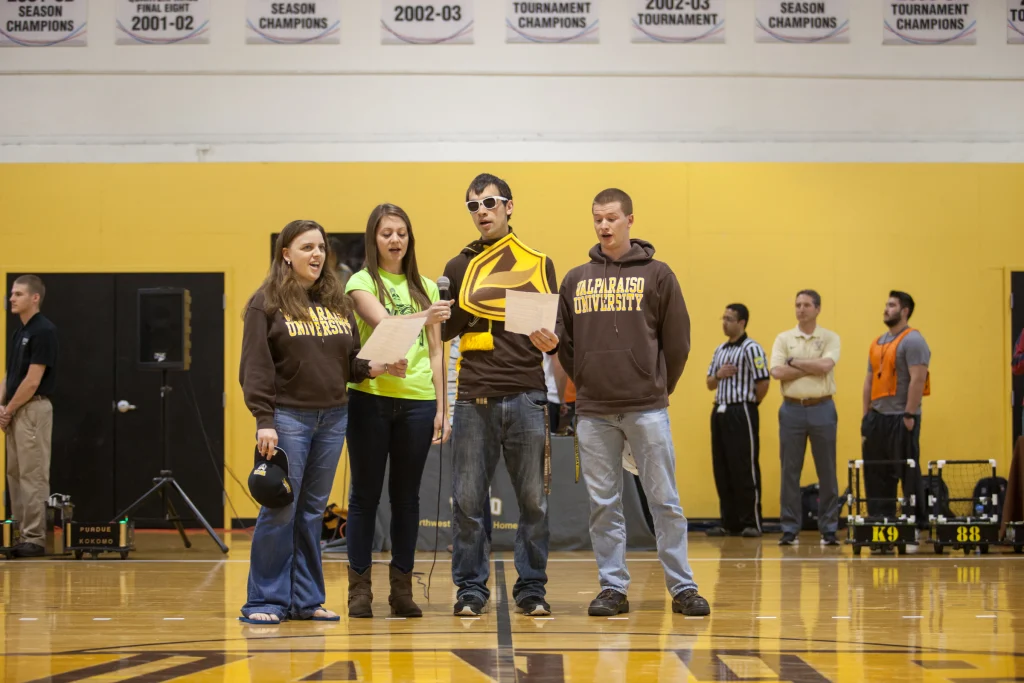 Four people stand on a gym floor, reading from papers, with Valpo and Purdue Kokomo banners and ROBOT FOOTBALL robots visible in the background.