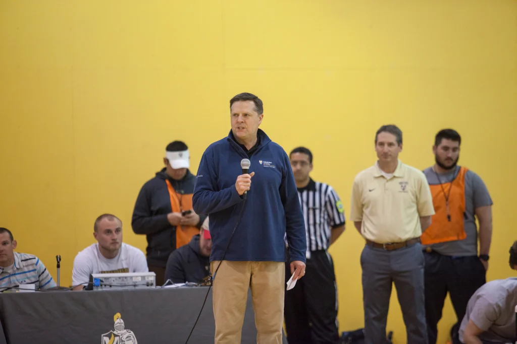 Man in a blue jacket speaks into a microphone in a gym, with Valpo staff and ROBOT FOOTBALL officials from Purdue Kokomo standing behind him.