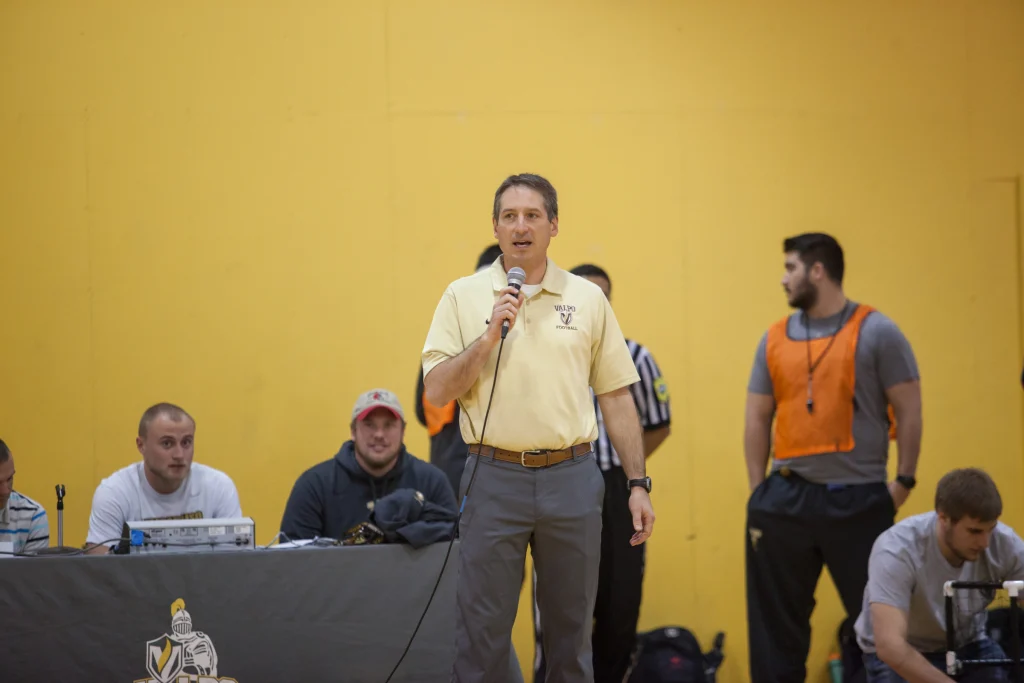 Man speaking into microphone in gym at Purdue Kokomo; others sit and stand nearby against a yellow wall. Robot football enthusiasts from Valpo join the event.