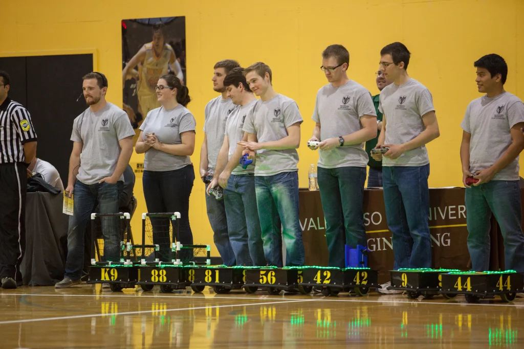A group of students in matching gray shirts stand behind their ROBOT FOOTBALL robots in a gym during a competition at Purdue Kokomo, ready to take on Valpo.
