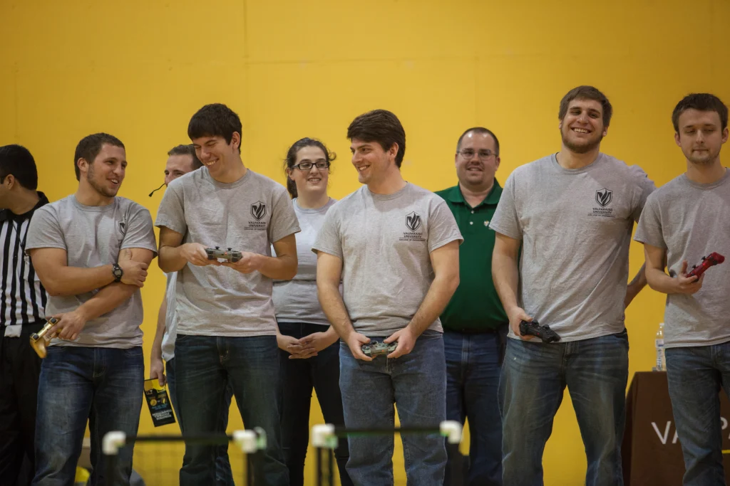 A group of people in gray shirts smiling and standing in front of a yellow wall at a Valpo robot football event.