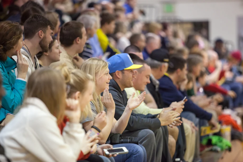 Crowd of people sitting in bleachers at an indoor event, some clapping and some looking at their phones during a Purdue Kokomo vs. Valpo ROBOT FOOTBALL match.