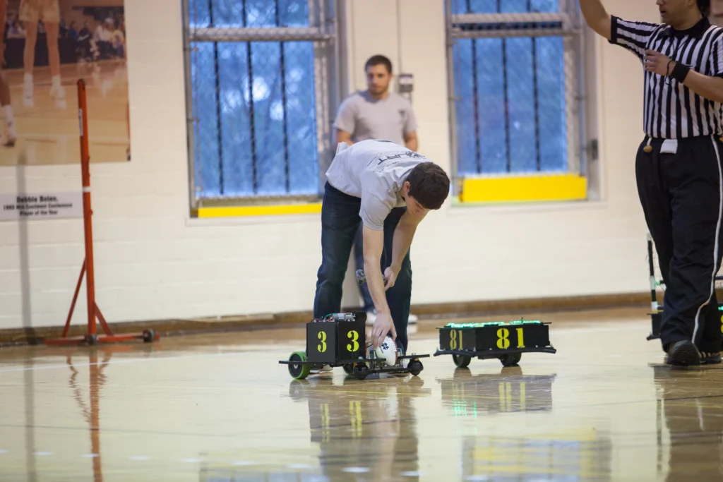 Person adjusting a small race car labeled "33" on a gym floor during a ROBOT FOOTBALL event; another car labeled "81" sits nearby, showcasing competition between Valpo and Purdue Kokomo.