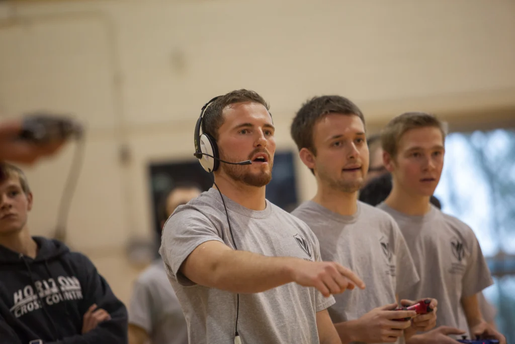 Three young men in gray shirts, one wearing a headset and pointing, focus intently indoors during a Purdue Kokomo ROBOT FOOTBALL event.
