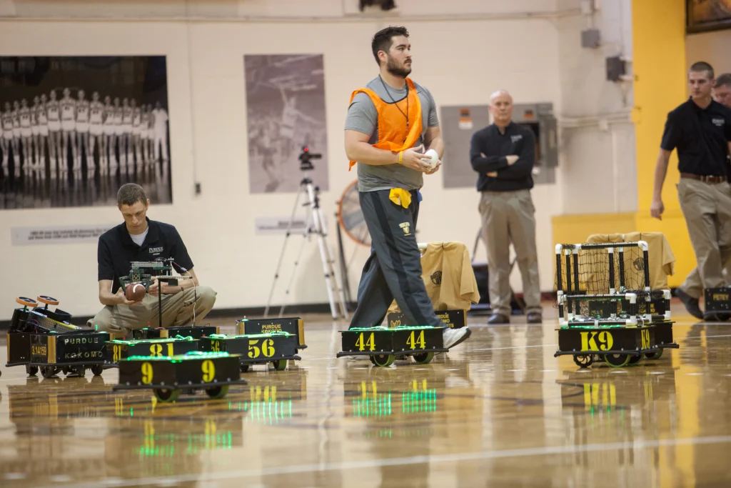 A man in an orange vest walks past several small, numbered robotic vehicles on a gym floor during a Purdue Kokomo vs. Valpo ROBOT FOOTBALL match.