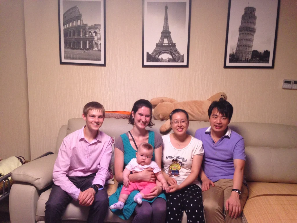 Five adults and a baby, including Aura Lee Harper-Smith, sit on a couch in a living room with travel photos adorning the wall behind them.