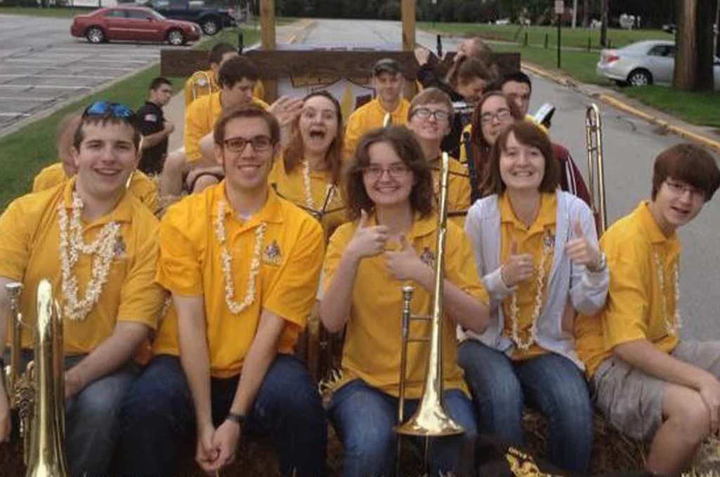A group of smiling students in yellow shirts with trombones pose together outdoors, giving thumbs up, showing their excitement for the upcoming Cody Johnson concert.