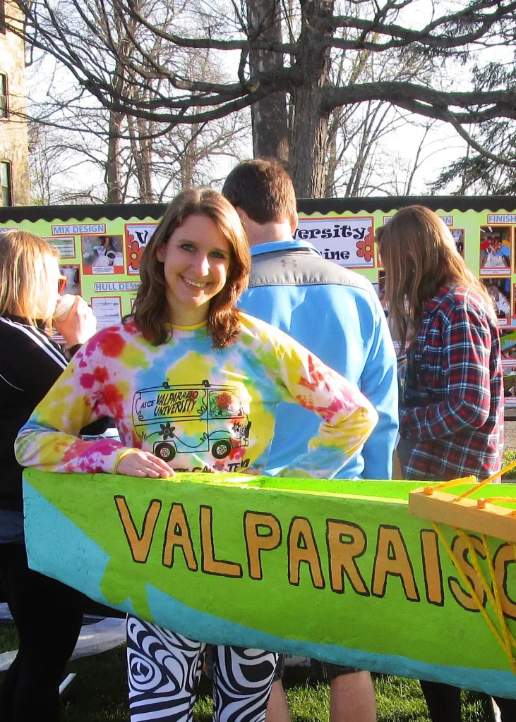 Young woman Eileen Carmignani in a tie-dye shirt smiles beside a green boat labeled "Valparaiso" at an outdoor event.
